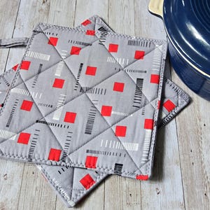 Könnte beinhalten: Ein quadratischer, gesteppter Topflappen aus grauem Stoff mit einem geometrischen Muster aus kleinen roten Quadraten sowie dünnen schwarzen und weißen gestrichelten Linien. Der Stoff ist rautenförmig gesteppt und hat eine Stoffschlaufe an der Ecke.
