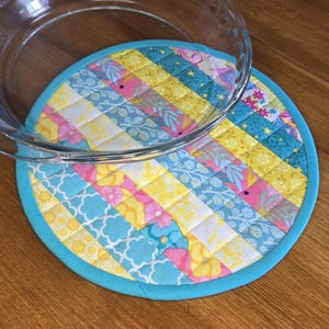 Könnte beinhalten: Ein runder, gesteppter Stoffuntersetzer mit einem Patchwork-Design aus gelben, rosafarbenen und hellblauen Blumen- und Geometriemustern, eingefasst mit einem türkisblauen Rand, teilweise bedeckt von einer gläsernen Kuchenform.