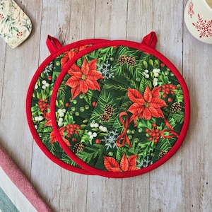 Peut inclure: Deux maniques rondes matelassées avec un fond vert foncé et un motif festif de fleurs de poinsettia rouges, de pommes de pin, de baies blanches et d'aiguilles de pin vertes. Chaque manique a une bordure en tissu rouge et une boucle.