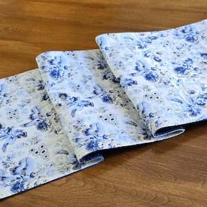 May include: A folded, quilted table runner featuring a floral design in blue and white hues. The runner has a light blue border and a repeating pattern of stylized flowers and leaves. The fabric appears to be cotton or a similar material.