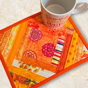 May include: A handmade orange quilted coaster with a floral design and a contrasting orange border. The coaster is placed under a white mug with the text "Pumpkin Kisses and Harvest Wishes".