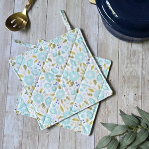Peut inclure: Deux maniques carrées matelassées avec un fond blanc et un motif répétitif de fleurs bleu clair, de feuilles vertes et de petits accents violets. Chaque manique possède une boucle en tissu dans le coin pour suspendre et des coutures en losange.