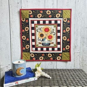 May include: A square fabric wall hanging featuring a central illustration of sunflowers in a vase with the word WELCOME on a banner. The design includes a black and white checkered border, a black floral sunflower print, and red trim.