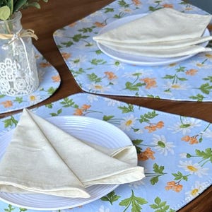 May include: A set of light blue placemats with a white daisy and orange flower pattern. Each placemat has a white plate with folded white napkins. A glass vase with white flowers is visible in the background.