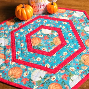 May include: A hexagonal quilted table mat featuring a teal background with a pattern of orange pumpkins, white daisies, and red leaves. The mat is accented with solid red fabric borders and straight-line quilting stitches throughout the design.