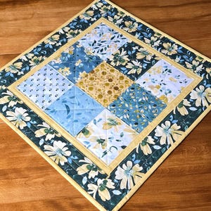 Peut inclure: Un set de table carré matelassé avec un motif floral. Le centre présente un patchwork de motifs floraux bleus, jaunes et blancs. Une bordure vert foncé avec des fleurs blanches et une bordure jaune entoure le centre. Le set de table mesure environ 30 cm de côté.