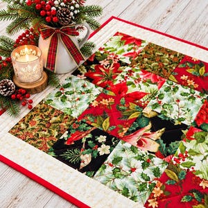 May include: A quilted table runner featuring a patchwork design of red poinsettias, green holly leaves, and red berries on black, white, and red backgrounds, bordered by a cream fabric with gold accents and a thin red trim.