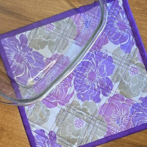 Könnte beinhalten: Ein quadratischer, gesteppter Topflappen mit Blumenmuster in Violett-, Lavendel- und Beigetönen auf weißem Grund, eingefasst mit einem violetten Stoffrand und diagonalen Steppnähten, teilweise bedeckt von einer klaren Glasschale.