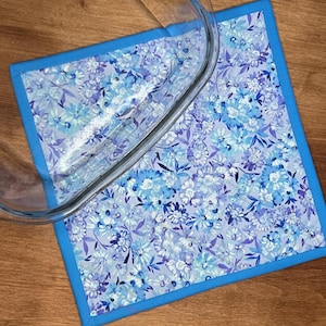 Könnte beinhalten: Ein quadratischer Topflappen aus Stoff mit einem hellblauen und violetten Blumenmuster und einem einfarbigen blauen Rand. Eine durchsichtige Glasauflaufform liegt teilweise auf dem Stoffpad und zeigt ihren geschwungenen Rand und Oberfläche.