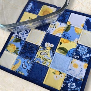 Peut inclure: Un dessous de plat carré matelassé avec un motif patchwork. Les carrés présentent des motifs floraux et cachemire dans des tons de bleu et de jaune, avec une bordure bleu foncé. Un plat en verre transparent repose sur le dessous de plat.