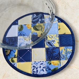 May include: A circular quilted hot pad with a patchwork design. It features squares of blue, yellow, and white fabric with floral and paisley patterns. A dark blue border surrounds the pad. A clear glass bowl rests on top.