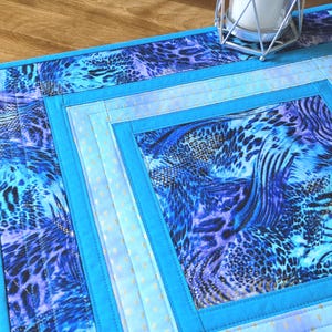 May include: A quilted table runner with a vibrant blue and purple animal print design. The runner features a square pattern with a light blue border and accents. The fabric has a shimmering effect, adding a touch of elegance to the home decor.
