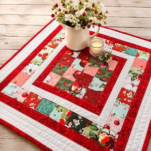 May include: A square quilted table topper featuring a patchwork design of red apples, green leaves, and white blossoms. The border consists of alternating red and patterned fabric squares, framed by white and solid red fabric strips.