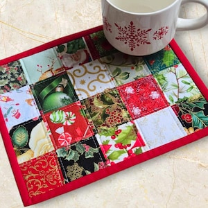 May include: A Christmas-themed quilted coaster with a patchwork design. The coaster features squares of fabric with festive patterns, including ornaments, holly, and snowflakes. A red border frames the coaster, and a white mug with a red snowflake design sits on top.