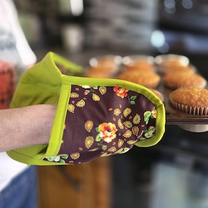 May include: An oven mitt featuring a solid lime green fabric on the interior and a dark brown exterior panel decorated with a repeating pattern of orange and yellow roses and small gold paisley shapes, finished with lime green fabric trim.