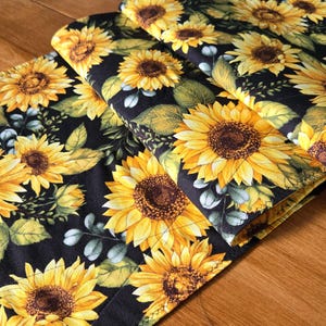 May include: Close-up of a floral fabric with large yellow sunflowers, brown centres, and green leaves on a black background. The fabric is folded, showing the repeating pattern. Edges are stitched with yellow thread.