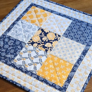 May include: A quilted table runner with a patchwork design featuring blue and yellow floral patterns. The runner has a white border with a blue trim.