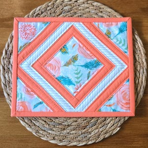 Peut inclure: Un dessous de plat matelassé carré avec un motif central en losange entouré de bordures concentriques. Le tissu présente un fond bleu clair avec des fleurs pêche et des plumes bleu sarcelle, rehaussé par des bandes de tissu corail uni.