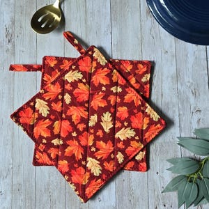 May include: Two square quilted pot holders featuring a repeating pattern of autumn maple and oak leaves in shades of red, orange, and yellow on a dark maroon background. Each pot holder includes a small fabric loop for hanging storage.