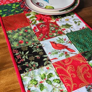 May include: A festive patchwork table runner with a red border. The runner features squares with Christmas patterns, including holly, poinsettias, and a cardinal bird. A white plate with a Christmas ornament design sits on top.