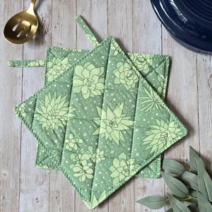 May include: Two square quilted pot holders featuring a light green fabric with a repeating pattern of various succulent plants, small white dots, and tiny star shapes. Each pot holder has a fabric loop attached to one corner for easy hanging storage.