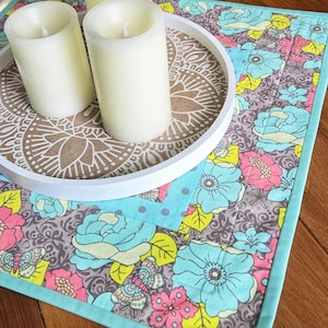 May include: A white-rimmed tray with two ivory candles rests on a floral placemat. The tray's centre has a brown mandala design. The placemat features large blue, pink, and yellow flowers. The candles are approximately 15 cm tall.