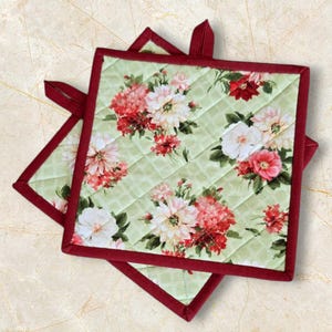May include: Two green quilted pot holders with a floral pattern and a burgundy border. The pot holders are made of fabric and have a quilted design.