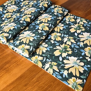 May include: A quilted table runner featuring a dark green background with a repeating pattern of yellow flowers and small blue leaves. The fabric has visible stitched lines forming a grid pattern across the surface of the floral textile.