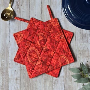 Peut inclure: Deux maniques matelassées en forme d'étoile dans des tons de rouge et d'orange avec un motif floral et géométrique complexe et répétitif. Chaque manique comporte une boucle en tissu au sommet pour la suspendre et des coutures diagonales.