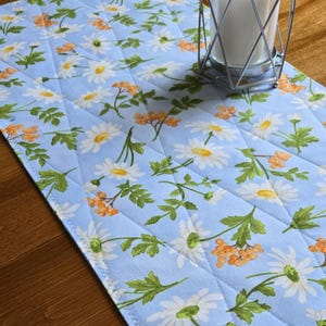 May include: A light blue quilted table runner with a daisy and berry pattern. The runner features white daisies with yellow centres, orange berries, and green leaves. A metal candle holder with a white candle sits on the runner.