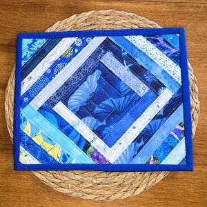 May include: A rectangular fabric quilt mat featuring a log cabin pattern in various shades of blue. The design consists of diagonal strips of fabric with diverse patterns, including floral and geometric motifs, finished with a solid blue border.
