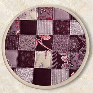 Könnte beinhalten: Ein runder, gesteppter Topflappen mit Patchwork-Design. Die Quadrate zeigen verschiedene florale und gemusterte Stoffe in Lila-, Burgunder- und Cremetönen. Der Rand ist mit einem hellbeigen Stoff verziert.