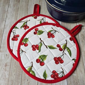 May include: Two round quilted pot holders featuring a white background with a repeating pattern of red cherries and green leaves. Each pot holder has a bright red fabric trim around the edge and a small red fabric loop for hanging storage.