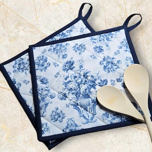 Könnte beinhalten: Zwei quadratische, gesteppte Topflappen mit blauem Blumenmuster und marineblauer Umrandung. Die Topflappen haben marineblaue Aufhängeschlaufen. Zwei Holzlöffel liegen neben den Topflappen.