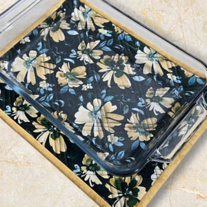 Peut inclure: Un plat à four en verre transparent repose sur un dessous de plat matelassé à motifs floraux. Le dessous de plat présente un fond noir avec des motifs de fleurs crème, vertes et bleues. Le dessous de plat a une bordure beige.