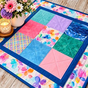 May include: A square quilted table topper featuring a nine-patch grid of varied fabric squares in teal, lavender, blue, pink, and floral patterns, framed by a solid dark blue border and an outer edge of colourful floral fabric with stitched details.