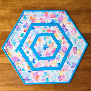 Peut inclure: Un dessous de plat hexagonal matelassé avec un motif floral aux tons rose, bleu, orange et violet. Le dessous de plat a une bordure turquoise vif et un motif hexagonal assorti au centre.