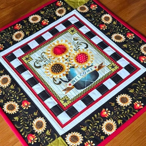 May include: A black and white checked fabric with a red border and a sunflower design in the centre. The design features a vase with sunflowers and the word "WELCOME" on a banner. The fabric is quilted and has a decorative border of sunflowers and vines.