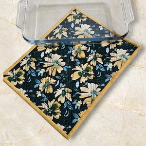 May include: A quilted, square hot pad with a floral pattern in cream, green, and blue on a dark background. The hot pad has a gold border and is shown with a glass baking dish.