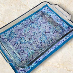 May include: A rectangular glass baking dish sits on a floral quilted pot holder. The pot holder has a blue and purple floral pattern with a turquoise border. The dish has handles. The pot holder is approximately 20 cm x 30 cm.