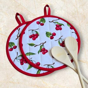 May include: Two round white and red pot holders with a cherry pattern and two wooden spoons. The pot holders have a red trim and a loop for hanging. The cherry pattern includes red cherries and green leaves.