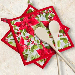 May include: Two red and white quilted pot holders with a poinsettia and holly pattern. Two wooden spoons are resting on top of the pot holders.