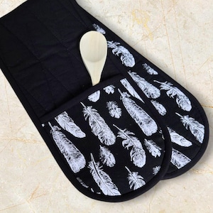 May include: A pair of black oven mitts with a white feather pattern. The mitts have a pocket on the side to hold a wooden spoon.