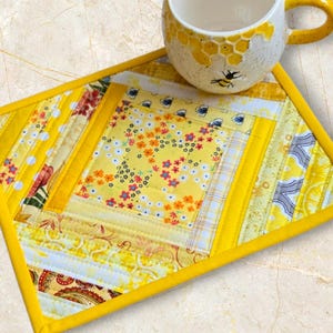 May include: A handmade yellow quilted placemat with floral and patterned fabrics. A yellow mug with a bee design sits on the placemat. The mug has a honeycomb pattern and a yellow handle.