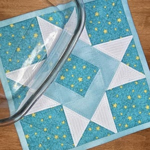 Könnte beinhalten: Ein quadratischer, gesteppter Topflappen aus blaugrünem Stoff mit gelbem Sternenmuster und weißen sternförmigen Akzenten. Der Stoff ist mit einem geometrischen Sternmuster abgesteppt und teilweise von einer Glasschale verdeckt.