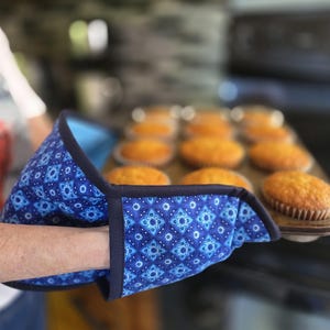 May include: A blue oven mitt with a repeating geometric pattern in shades of light and dark blue, featuring a dark blue fabric trim along the edges. The mitt is shown holding a metal baking tray containing several golden-brown baked muffins.