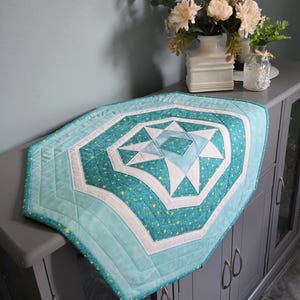 May include: A quilted table runner featuring a central star pattern in shades of teal, light blue, and white. The fabric displays a repeating pattern of small yellow stars on a teal background, accented by solid white and light blue geometric borders.