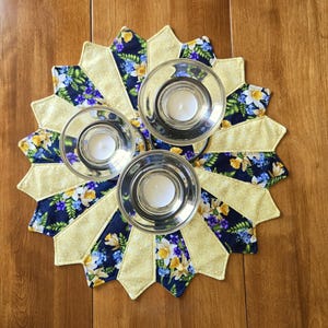 Peut inclure: Un tapis en tissu circulaire présentant un motif en étoile avec des panneaux alternés jaune clair et bleu foncé à imprimé floral de jonquilles jaunes et de fleurs violettes. Trois bougeoirs en verre transparent sont posés dessus.