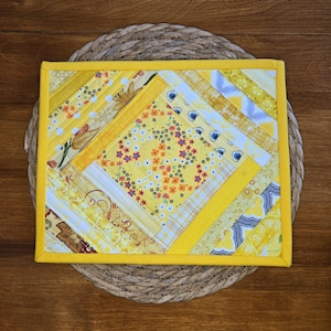 Könnte beinhalten: Ein quadratischer, gesteppter Topflappen mit einem Patchwork-Design aus verschiedenen gelben, weißen und grauen Stoffen. Das mittlere Quadrat zeigt ein kleines Blumenmuster, umgeben von diagonalen Stoffstreifen mit Punkten, Schnörkeln und botanischen Motiven.