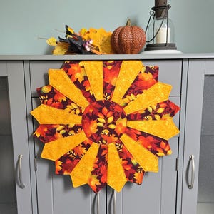 May include: A circular quilted fabric mat featuring a sunburst pattern of alternating golden yellow and dark maroon fabric segments, each decorated with a printed autumn leaf motif. The centre is a round patch of dark fabric with leaves.
